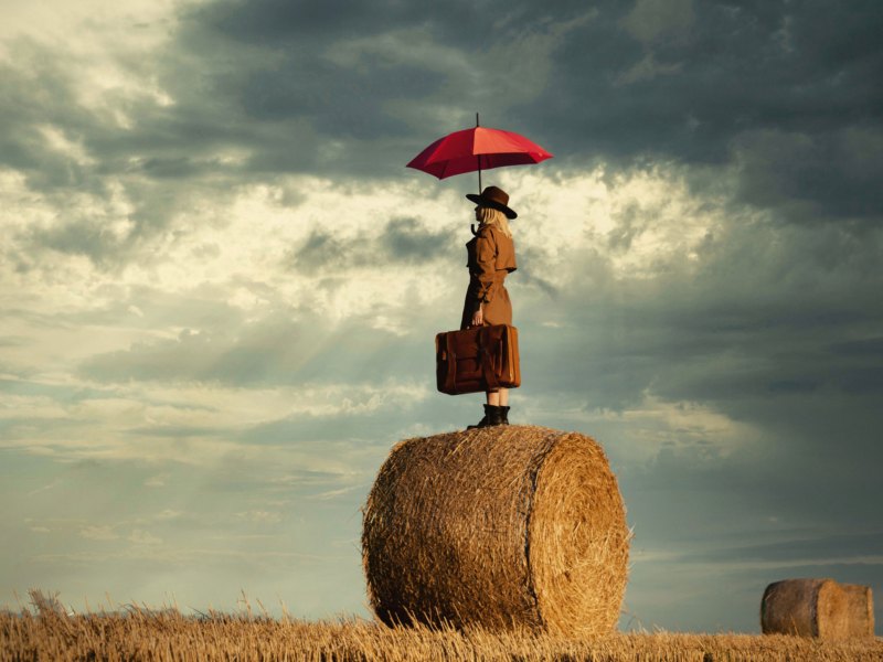 Frau mit Regenschirm und Aktenkoffer auf einem Strohballen auf einem Feld.