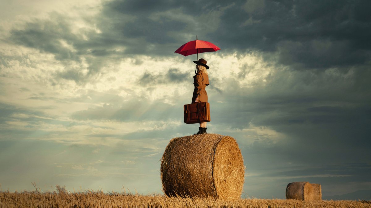 Frau mit Regenschirm und Aktenkoffer auf einem Strohballen auf einem Feld.