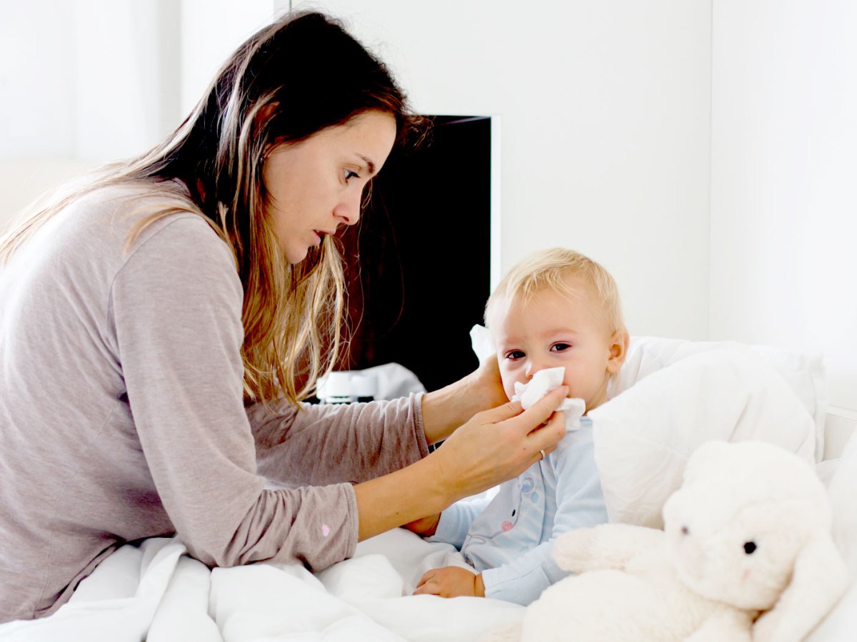 Mutter sitzt am Bett ihres ca. 2-jährigen Sohnes und wischt ihm die Nase ab.