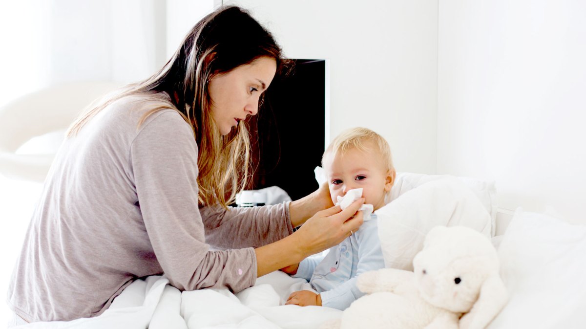 Mutter sitzt am Bett ihres ca. 2-jährigen Sohnes und wischt ihm die Nase ab.