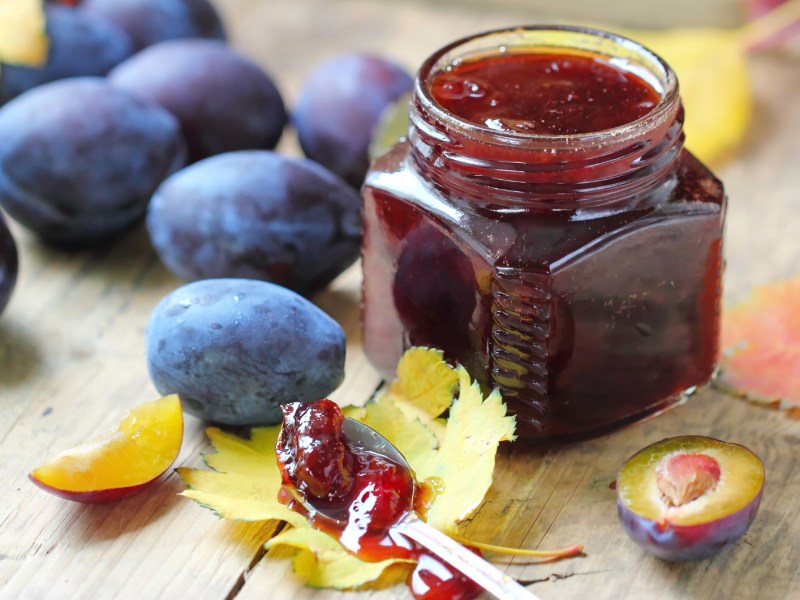 Pflaumenmarmelade in einem Weckglas, daneben liegen frische Pflaumen und Herbstblätter.