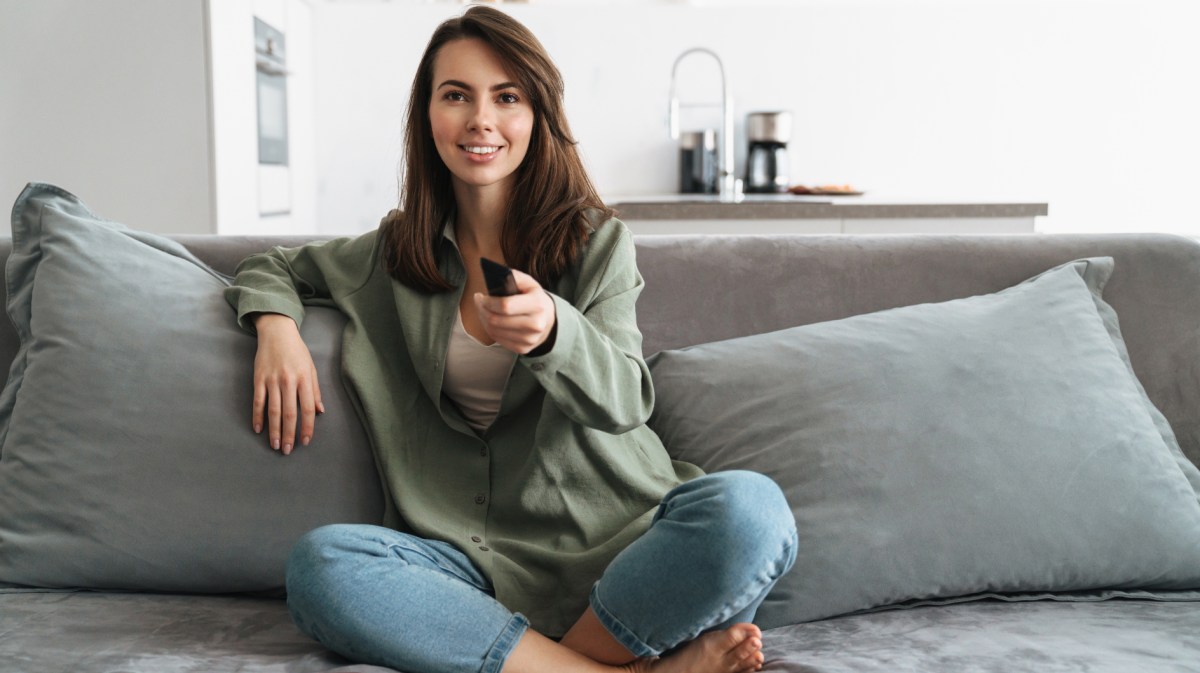 Brünette Frau sitzt auf grauem Sofa, die lächelt und hat Fernbedienung auf Kamera gerichtet.