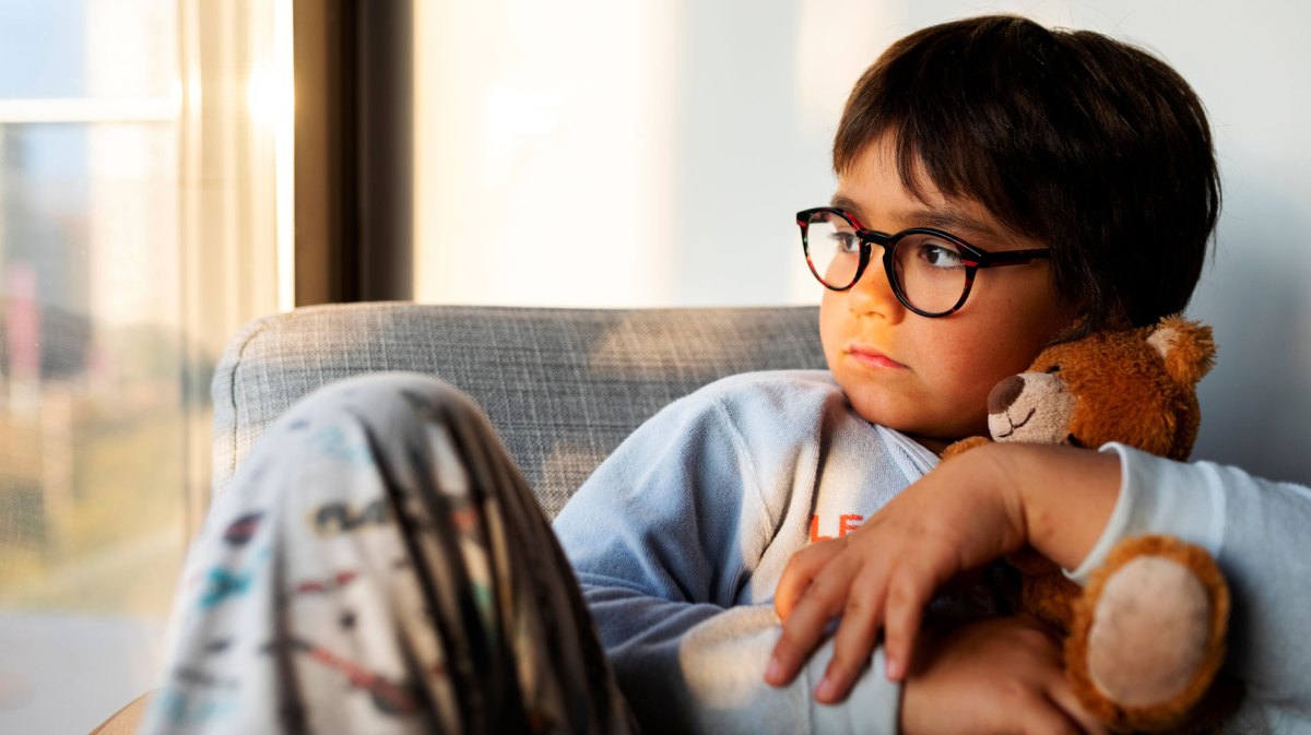 Porträt eines Jungen, ca. 10 Jahre alt, der mit einem Teddy im Arm melancholisch aus dem Fenster schaut.