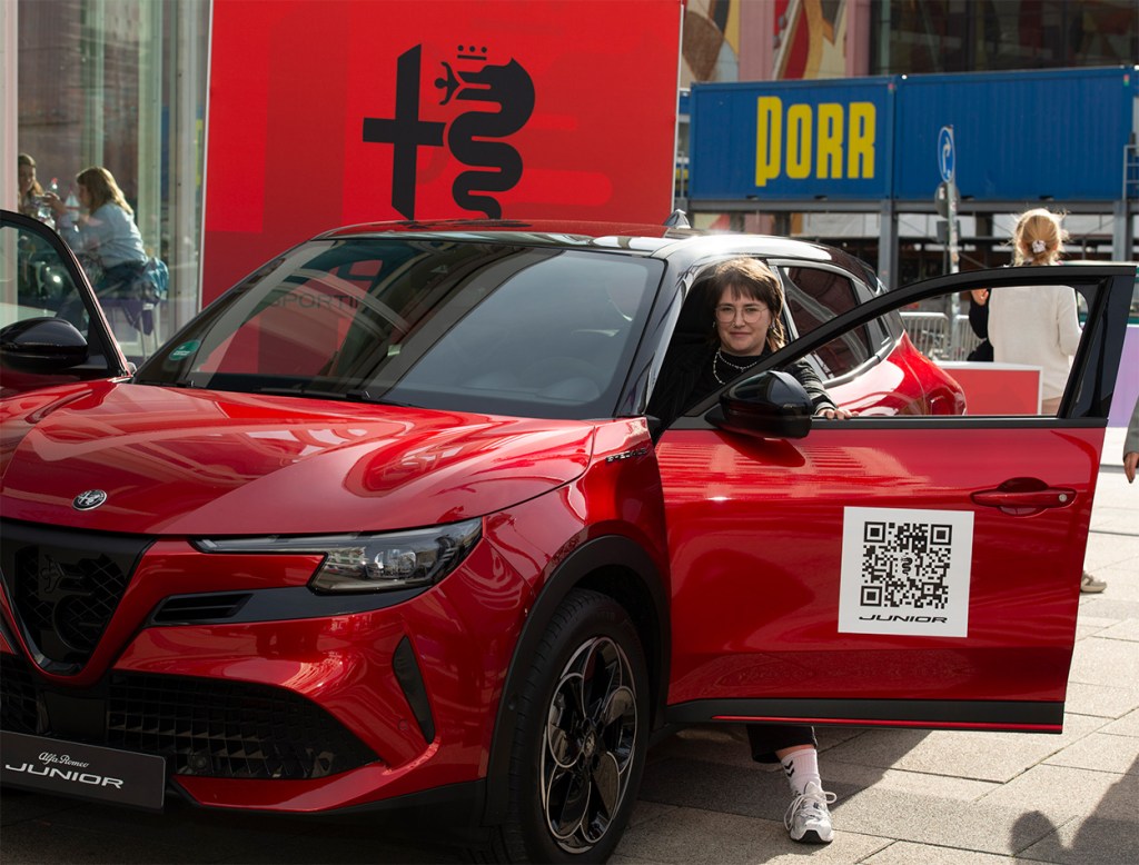 Eine junge Frau mit Brille sitzt lächelnd in einem roten Alfa Romeo, die Beifahrertür ist geöffnet, im Hintergrund eine Werbewand.