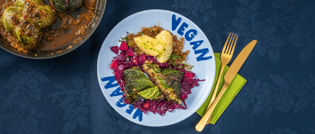 Gefüllte Kohlrouladen mit Bratensoße, Kartoffeln und Rotkraut auf einem weißen Teller mit "Vegan"-Schriftzug, daneben goldenes Besteck.