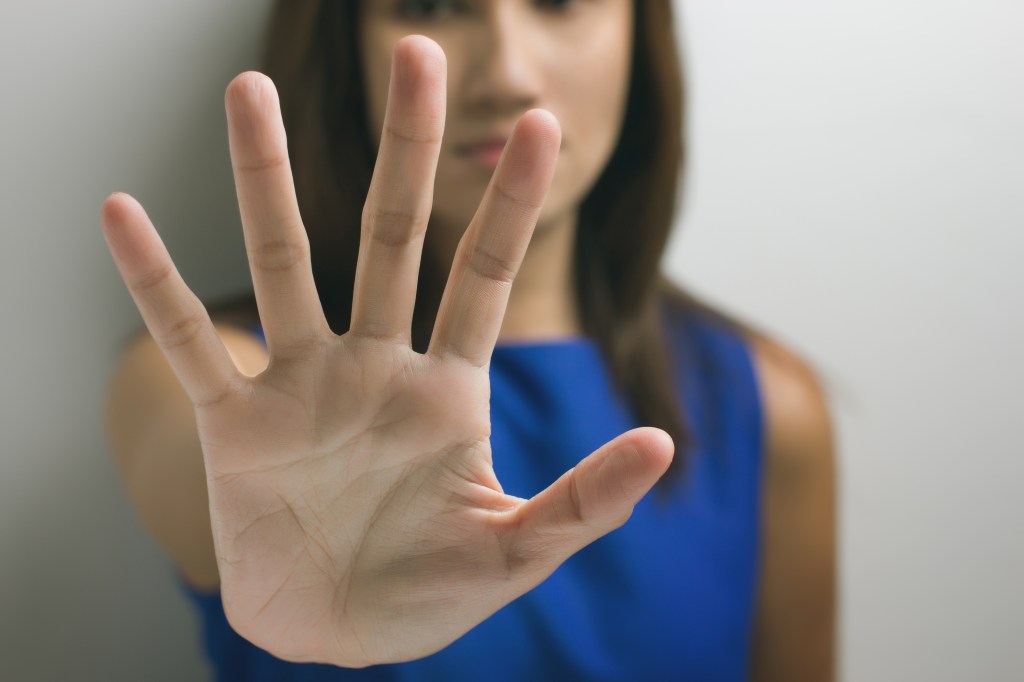 Junge Frau in blauem Kleid hält die offene Handfläche abweisend nach vorne, ihr Gesicht ist unscharf im Hintergrund.