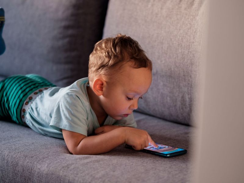 Kleinkind liegt auf dem Sofa und schaut auf ein Handy