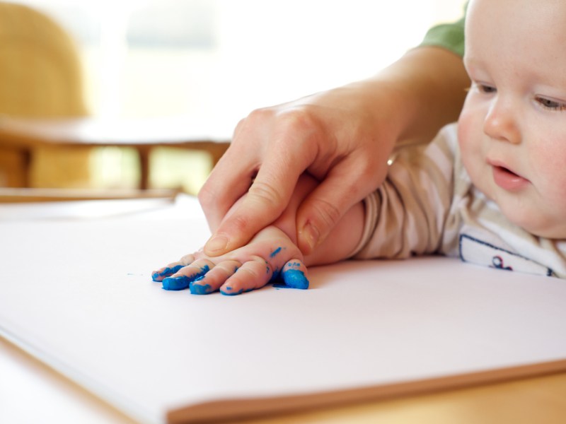 Ein Kleinkind macht mit Mamas Hilfe und Fingerfarbe einen Handabdruck auf einem Blatt Papier