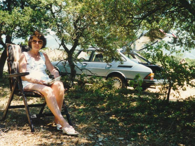 Bild aus den 80er Jahren zeigt eine Frau auf einem Campingstuhl, im Hintergrund ist ein altes Auto zu sehen.