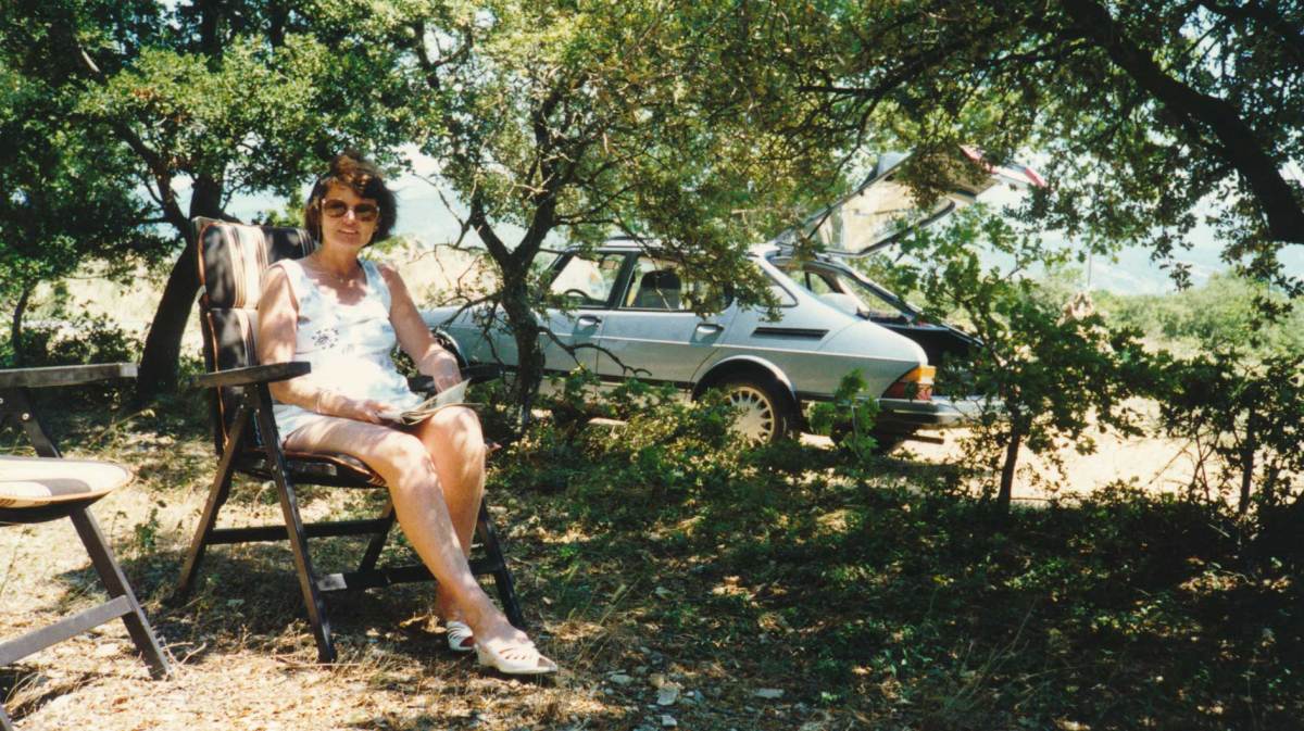 Bild aus den 80er Jahren zeigt eine Frau auf einem Campingstuhl, im Hintergrund ist ein altes Auto zu sehen.