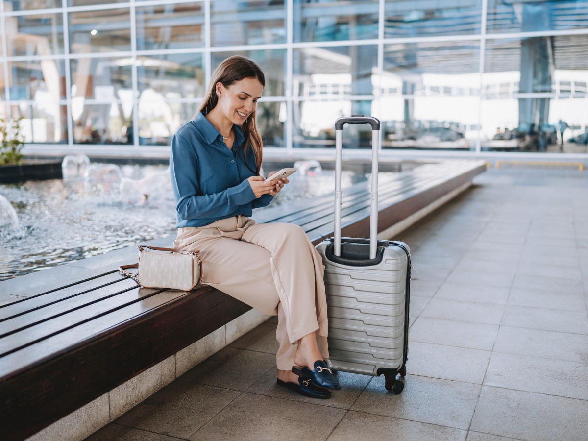 Frau sitzt am Flughafen schaut auf Handy, daneben steht ein heller Koffer