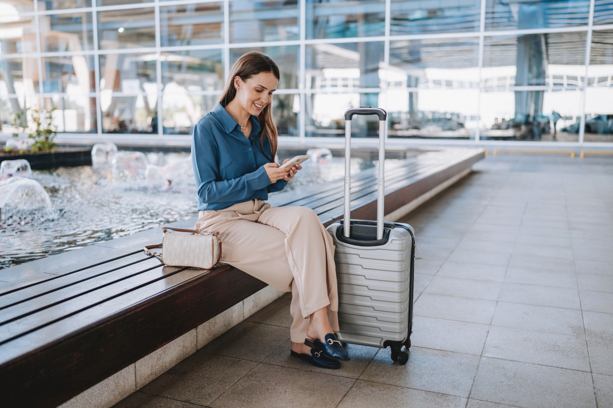 Frau sitzt am Flughafen schaut auf Handy, daneben steht ein heller Koffer