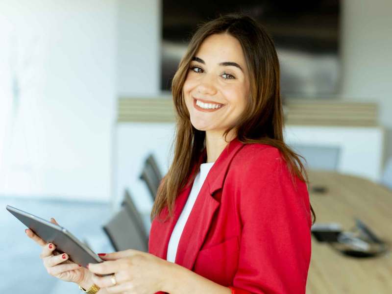 Junge Frau mit rotem Blazer steht im Büro und hält ein Tablet fest.