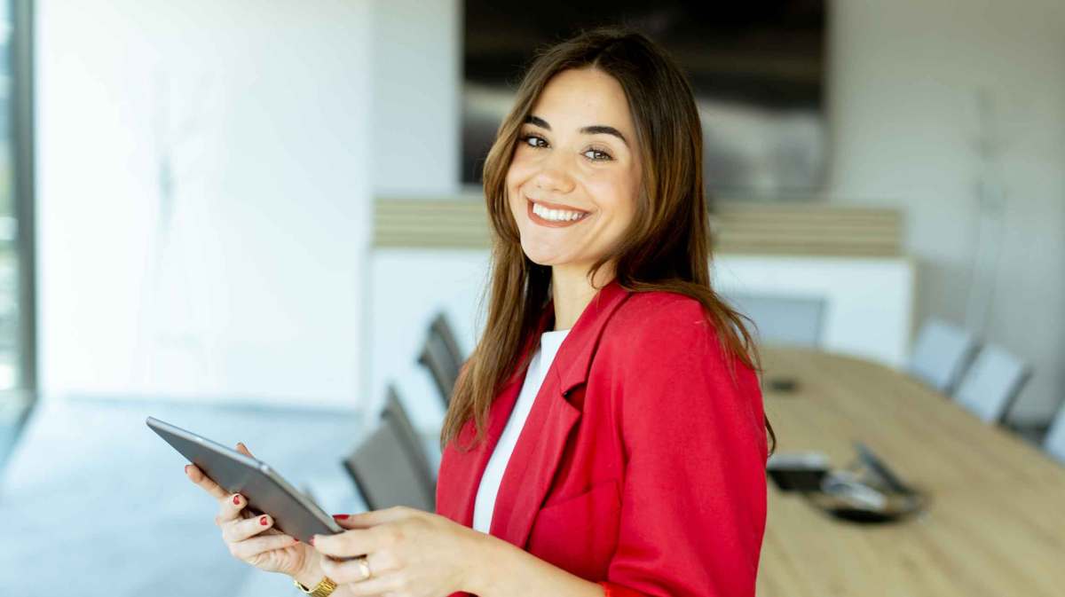 Junge Frau mit rotem Blazer steht im Büro und hält ein Tablet fest.