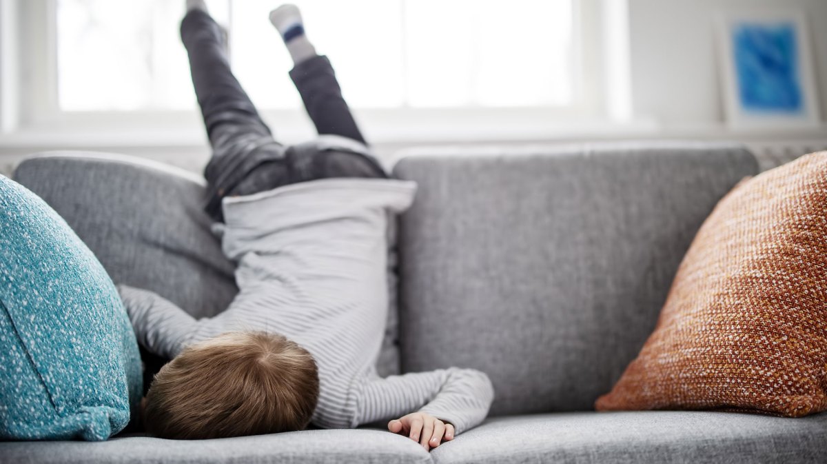 Junge hat sich über die Lehne des Sofas geworfen und liegt mit den Füßen oben und dem Gesicht auf den Sitzkissen gelangweilt herum.