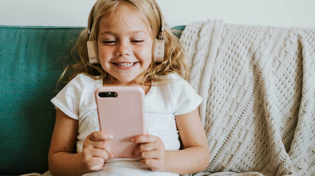 Mädchen, ca. 6 Jahre, sitzt mit Kopfhörern und Handy auf dem Sofa und schaut sich etwas auf dem kleinen Bildschirm an.