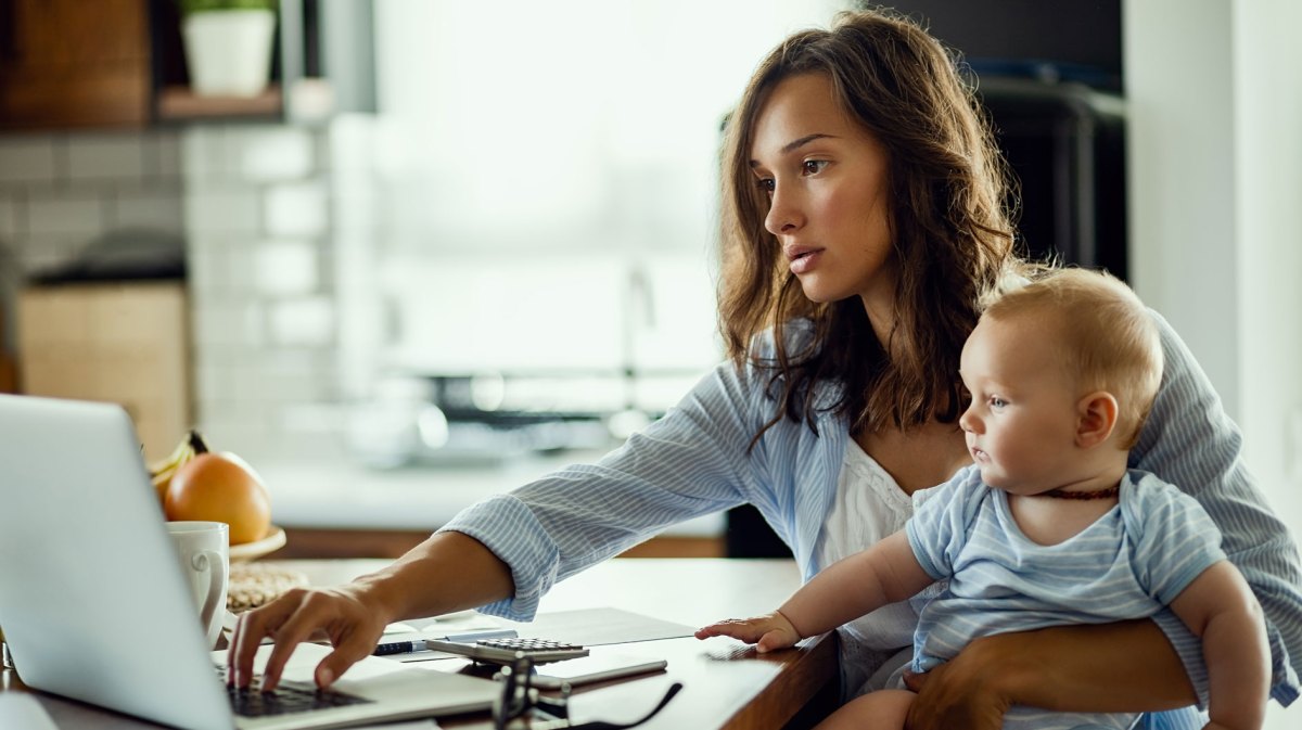 Junge Mutter sitzt mit Baby auf dem Schoß vor ihrem Laptop.