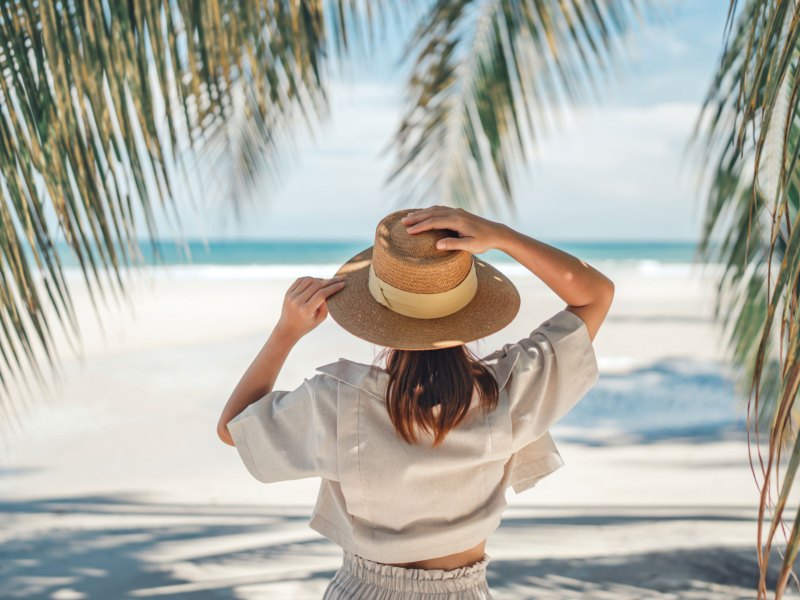 Frau mit Sonnenhut steht am Strand unter einer Palme und schaut aufs Meer