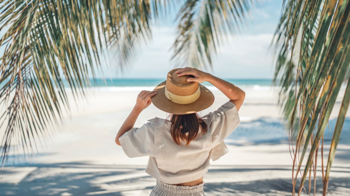 Frau mit Sonnenhut steht am Strand unter einer Palme und schaut aufs Meer