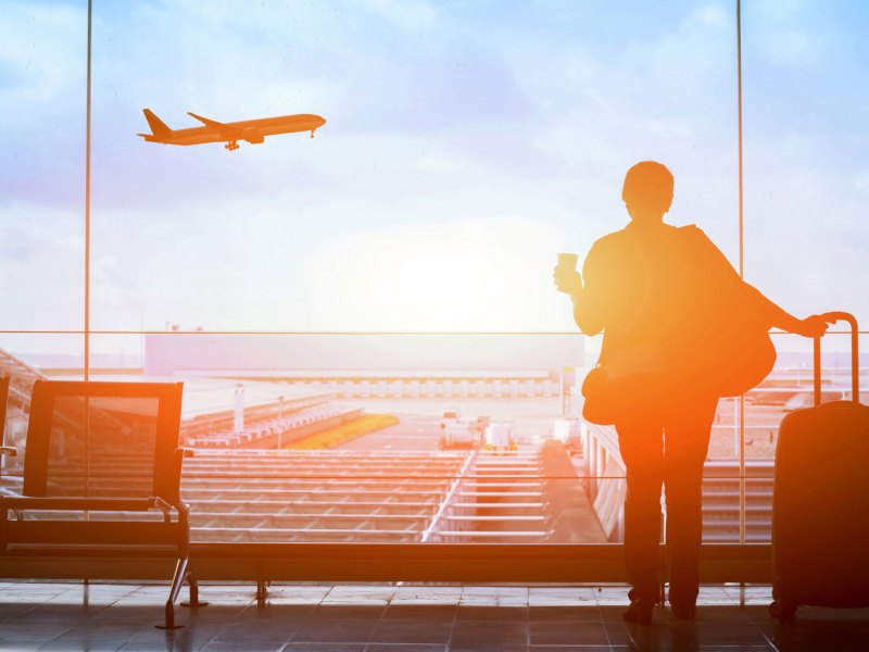 Frau mit Koffer in der Hand am Flughafen schaut raus auf startendes Flugzeug