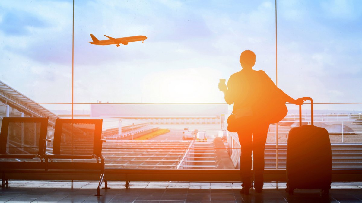 Frau mit Koffer in der Hand am Flughafen schaut raus auf startendes Flugzeug