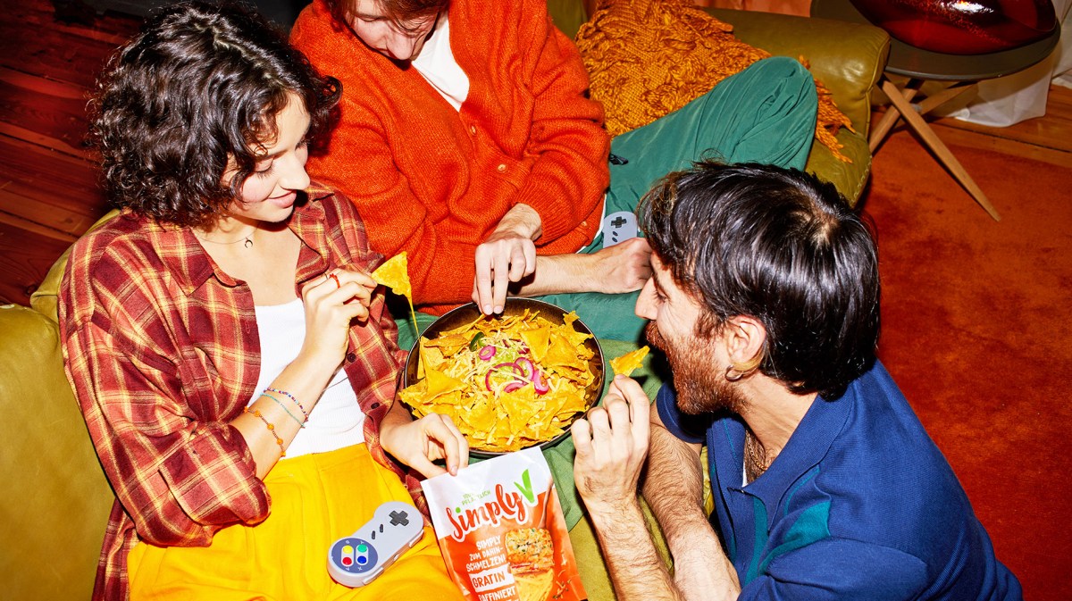 Drei junge Freunde in bunter Kleidung sitzen auf einem Sofa und teilen eine Schüssel Nachos mit Käse und Zwiebeln.