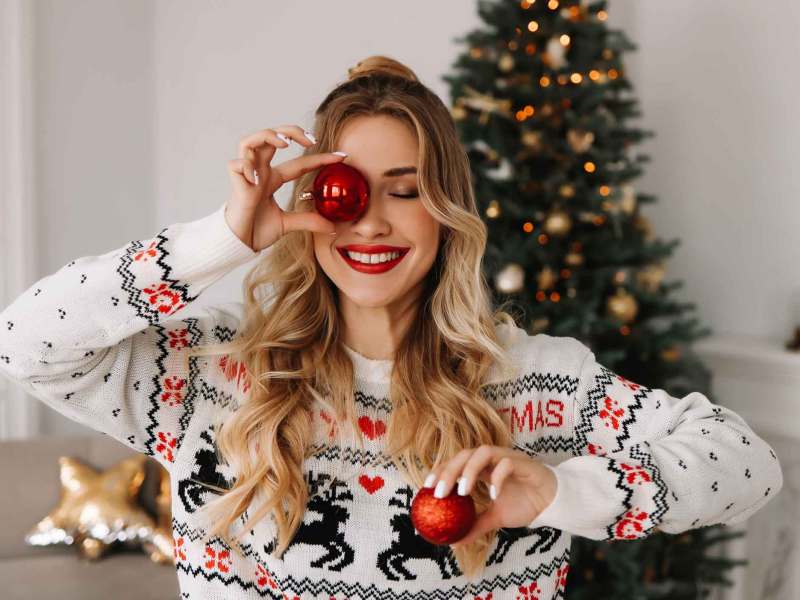 Frau mit langen Haaren hält zwei Christbaumkugeln in der Hand. Die eine hält sie sich vors Auge und lacht dabei. Im Hintergrund steht ein geschmückter Weihnachtsbaum.