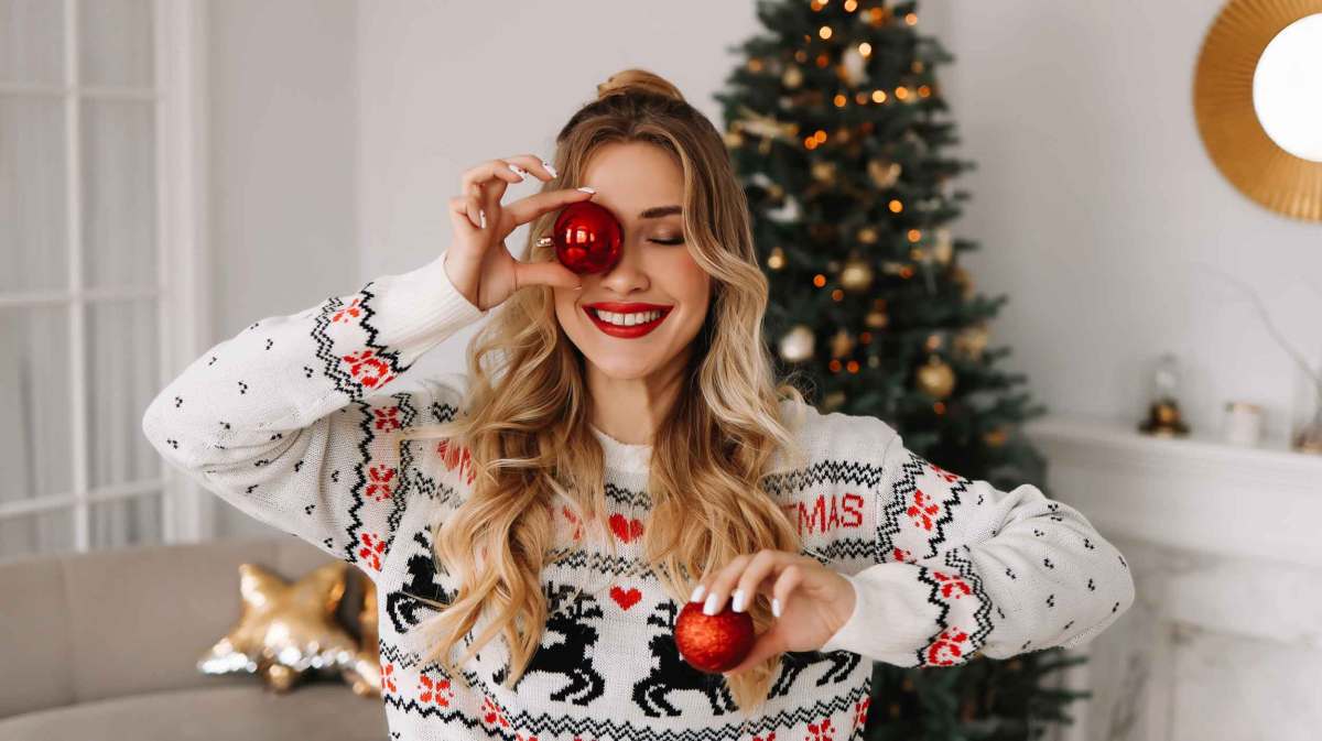 Frau mit langen Haaren hält zwei Christbaumkugeln in der Hand. Die eine hält sie sich vors Auge und lacht dabei. Im Hintergrund steht ein geschmückter Weihnachtsbaum.