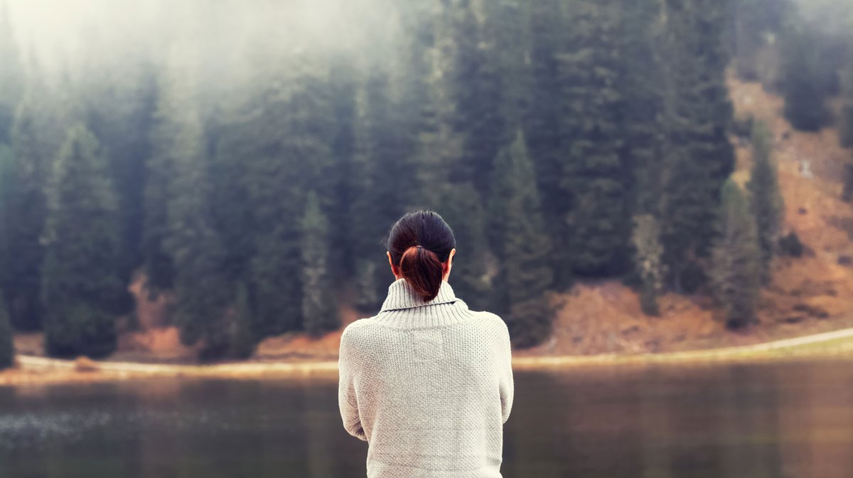 Frau steht an einem See mit Wald im Hintergrund