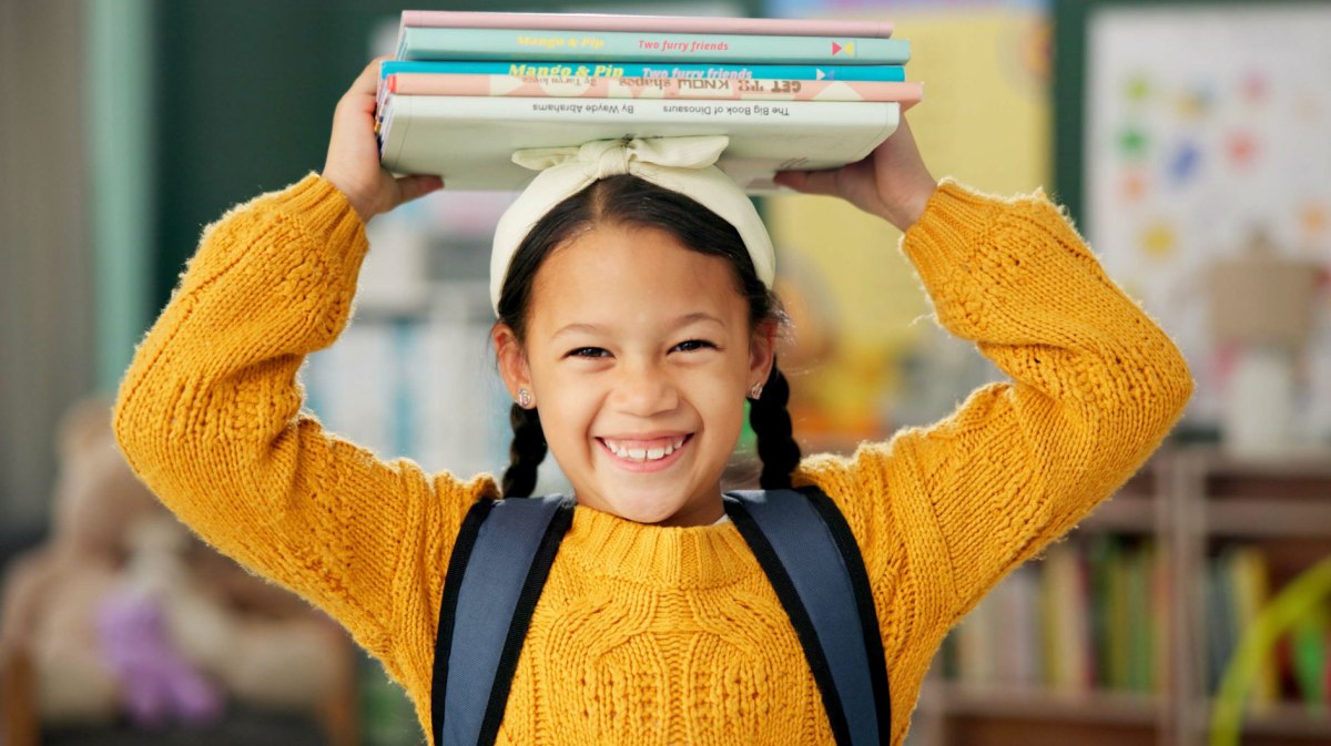 Lachendes Mädchen hält einen Stapel Bücher mit beiden Händen seitlich auf ihrem Kopf fest und steht in einem Klassenzimmer.