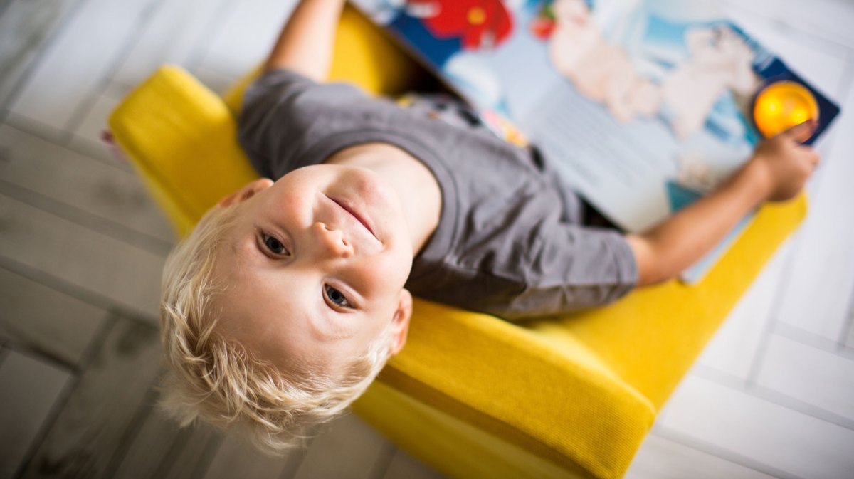 Junge, ca. 5 Jahre, sitzt auf einem gelben Kindersessel, hat ein Buch auf dem Schoß und schaut nach oben in die Kamera.