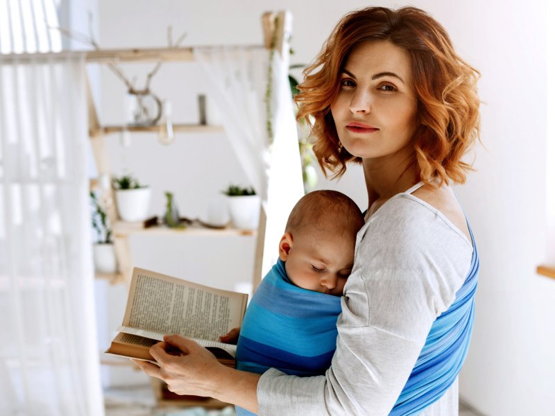 Porträt einer zufriedenen Mutter, die ihr Kind in einem Tragetuch vor der Brust trägt und ein Buch in den Händen hält.