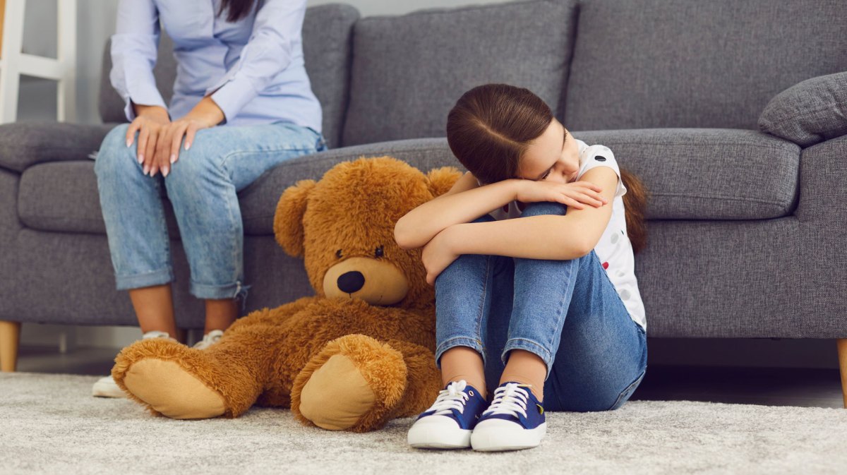 Mädchen sitzt traurig auf dem Boden, an ein Sofa gelehnt. Die Mutter sitzt im Hintergrund, ist nur angeschnitten zu sehen.