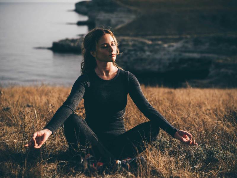 Junge Frau in schwarzem Hemd übt Yoga-Asanas auf hoher Felsenküste über dem Meer bei hellem Sonnenlicht und leichtem Wind in ruhigem Morgen