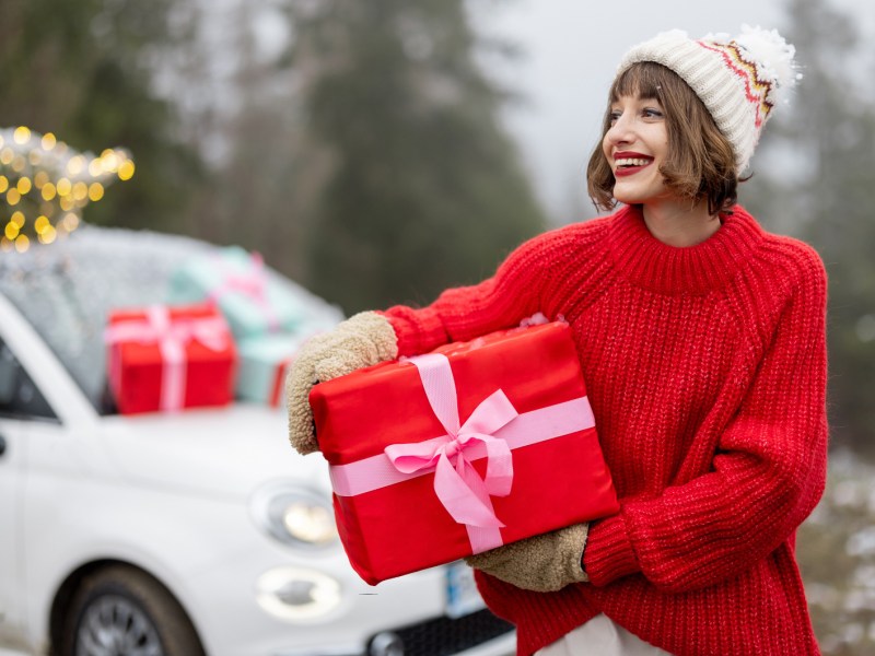 Frau mit kurzen Haaren hält Geschenk und steht vor weißem Auto.