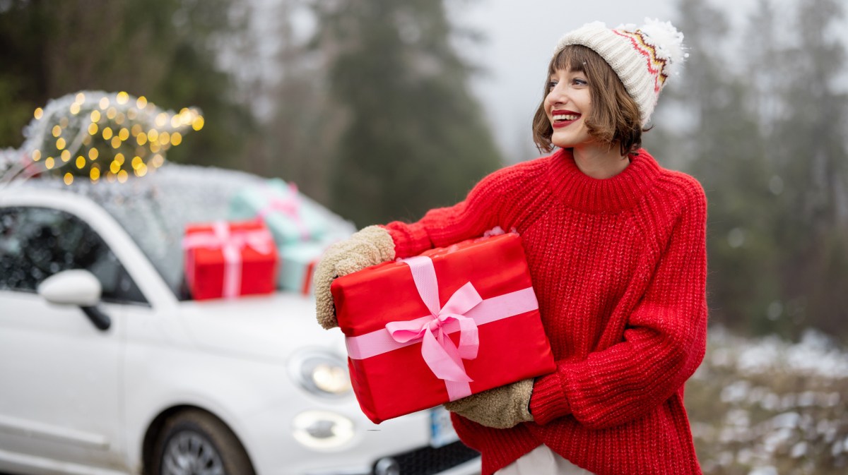 Frau mit kurzen Haaren hält Geschenk und steht vor weißem Auto.