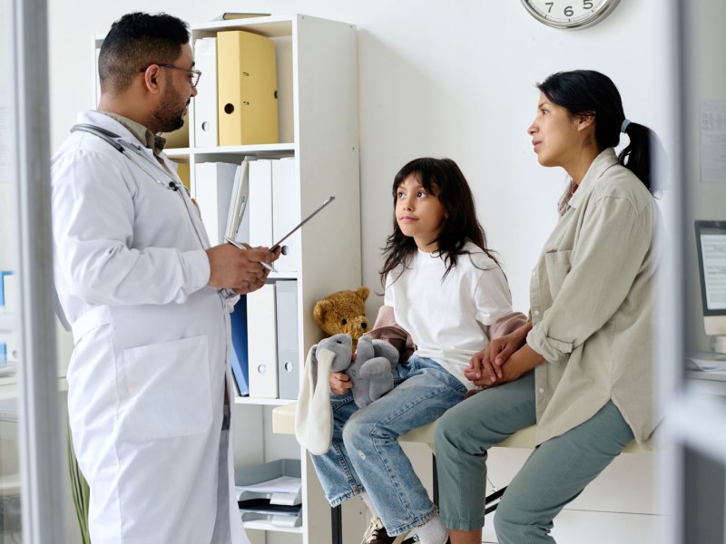 Mutter und Tochter sitzen im Behandlungszimmer auf einer Liege, ein Arzt steht vor ihnen und erklärt etwas.