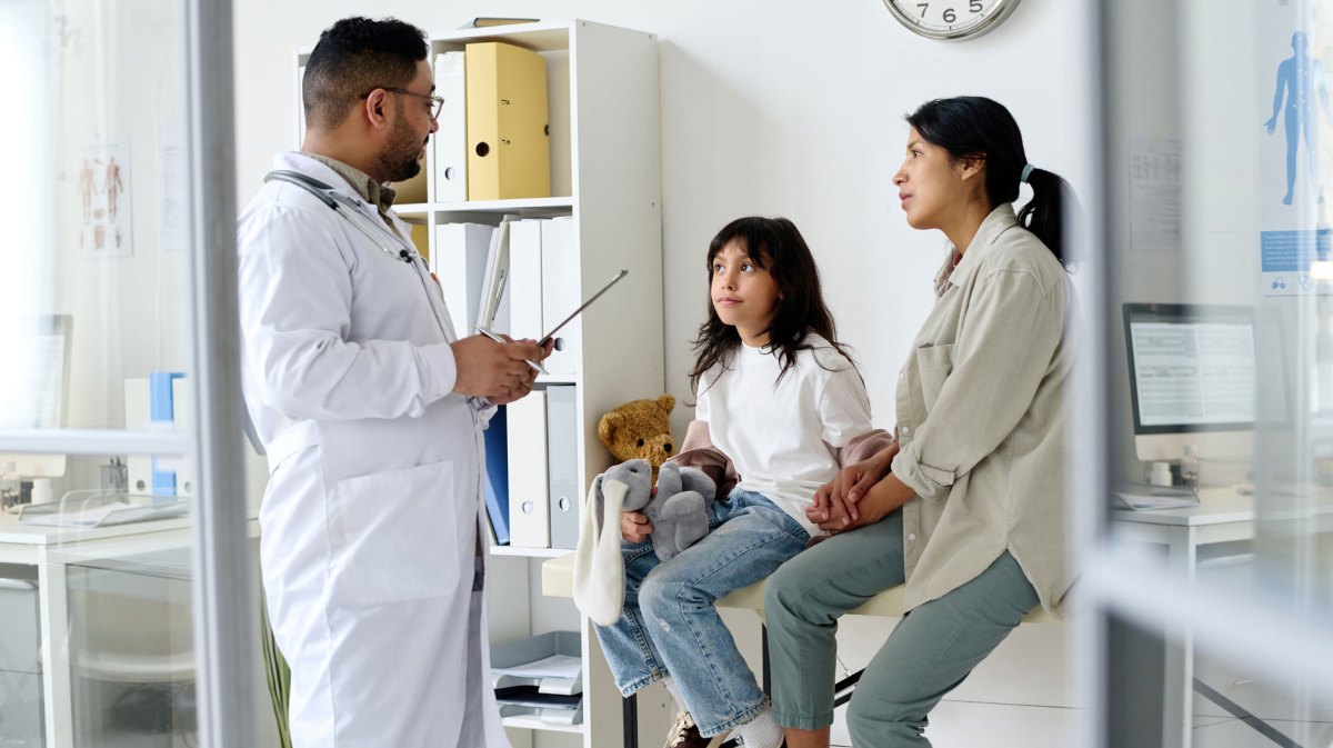 Mutter und Tochter sitzen im Behandlungszimmer auf einer Liege, ein Arzt steht vor ihnen und erklärt etwas.