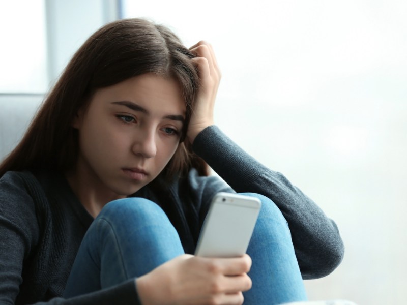 Junges Mädchen sitzt mit einem Handy nachdenklich vor dem Fenster