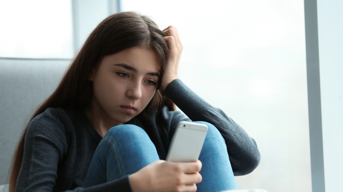 Junges Mädchen sitzt mit einem Handy nachdenklich vor dem Fenster