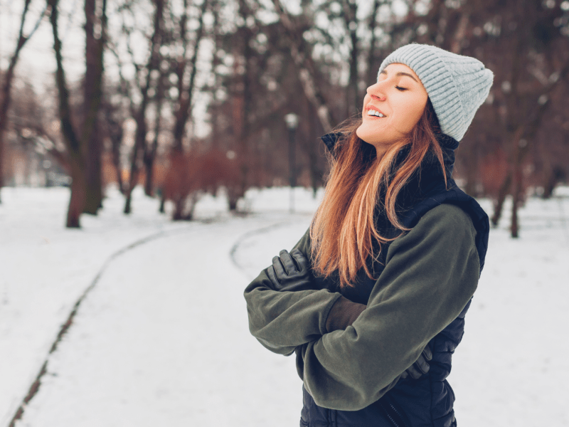 Junge Frau mit langen braunen Haaren und grauer Strickmütze steht lächelnd in einem verschneiten Park und genießt die Winterluft.