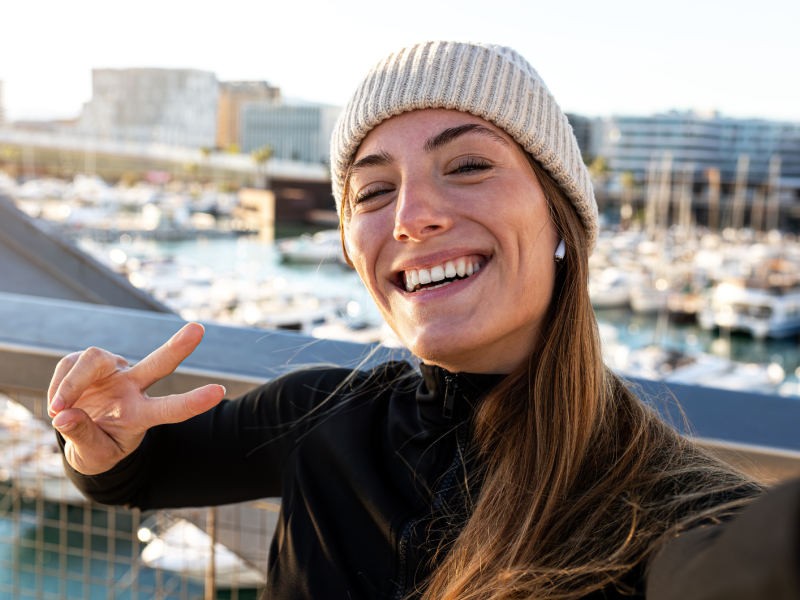 Fröhliche Frau in schwarzer Jacke und beige Strickmütze zeigt ein Peace-Zeichen, während sie ein Selfie vor einem Yachthafen aufnimmt.