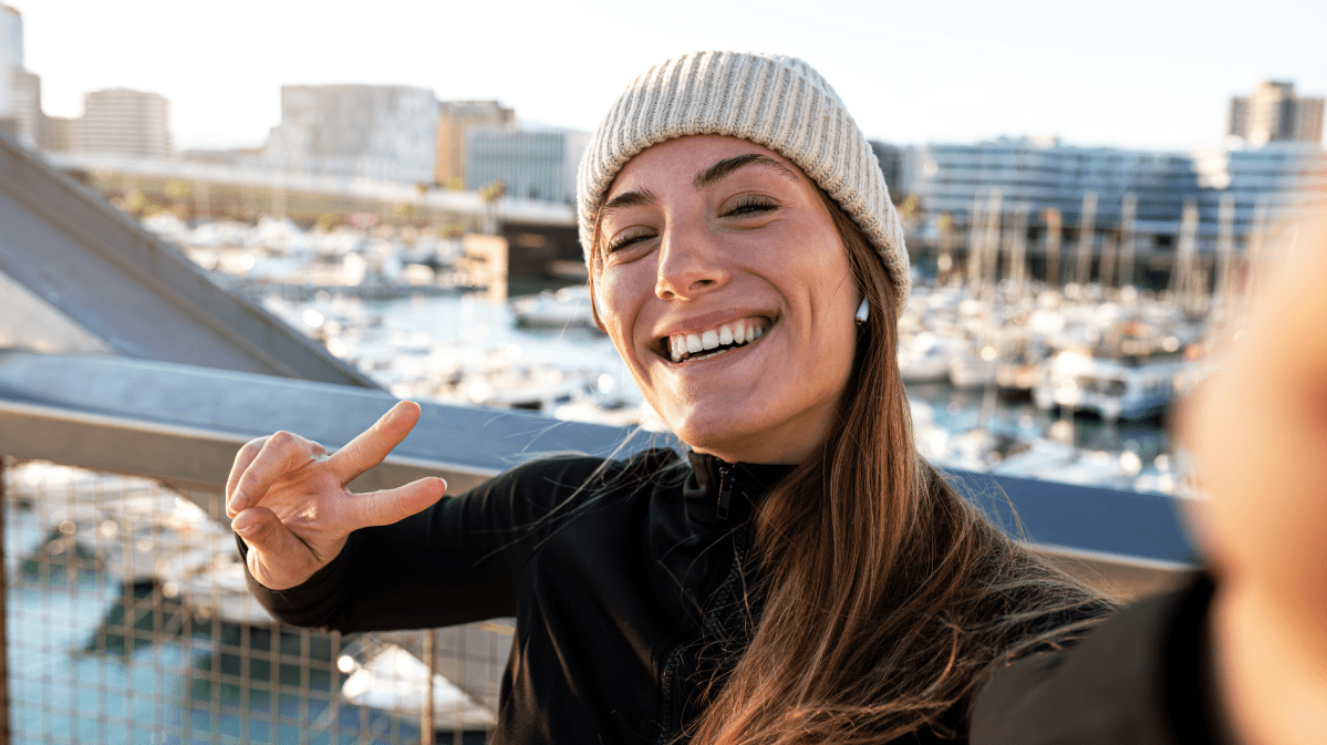 Fröhliche Frau in schwarzer Jacke und beige Strickmütze zeigt ein Peace-Zeichen, während sie ein Selfie vor einem Yachthafen aufnimmt.