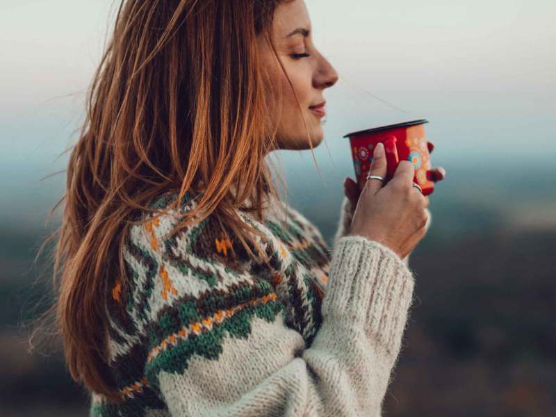 Frau mit Tasse und Strickpullover steht draußen vor einer Landschaft.