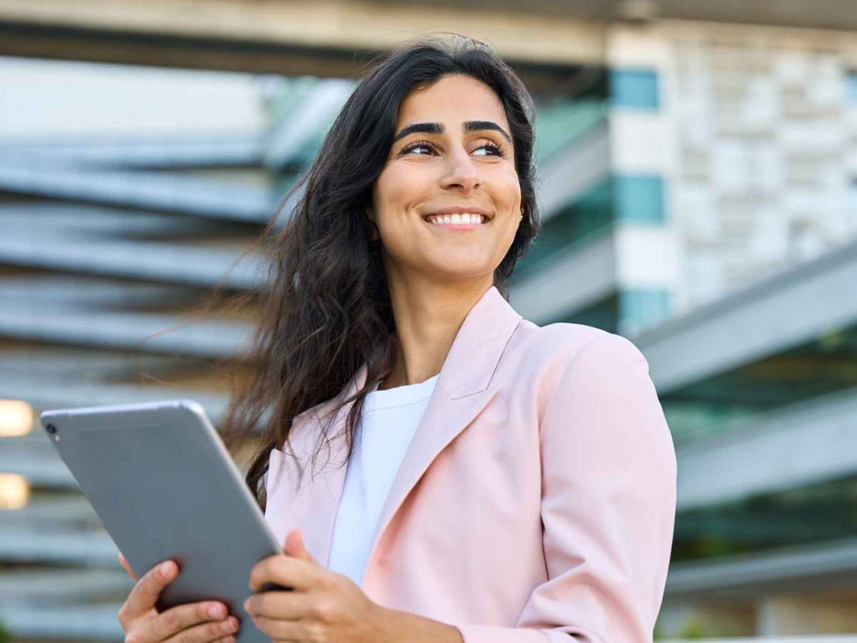 Frau mit rosa Blazer steht draußen vor einem Bürogebäude mit einem Tablet.