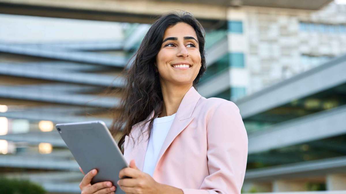 Frau mit rosa Blazer steht draußen vor einem Bürogebäude mit einem Tablet.