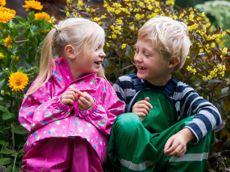 Zwei Kinder hocken an einem Herbsttag in Regensachen vor einem gelben Blumenbusch.