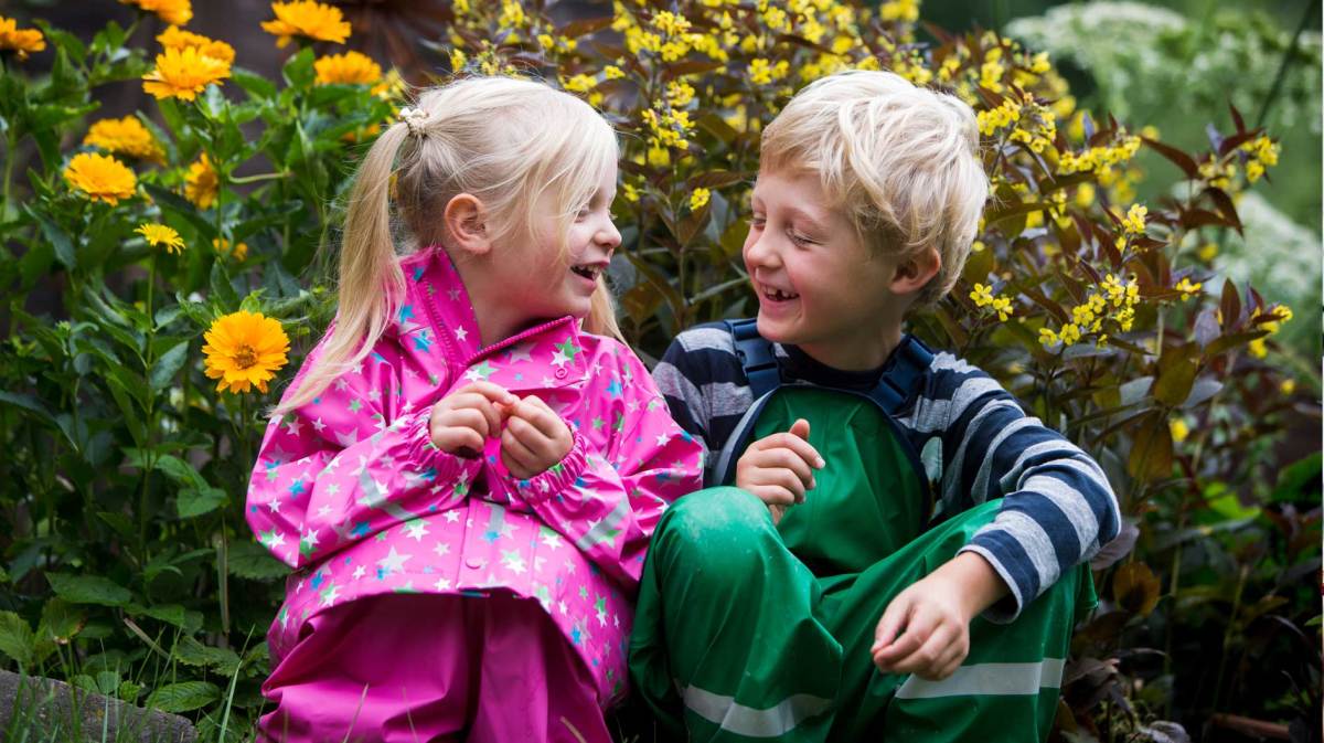 Zwei Kinder hocken an einem Herbsttag in Regensachen vor einem gelben Blumenbusch.