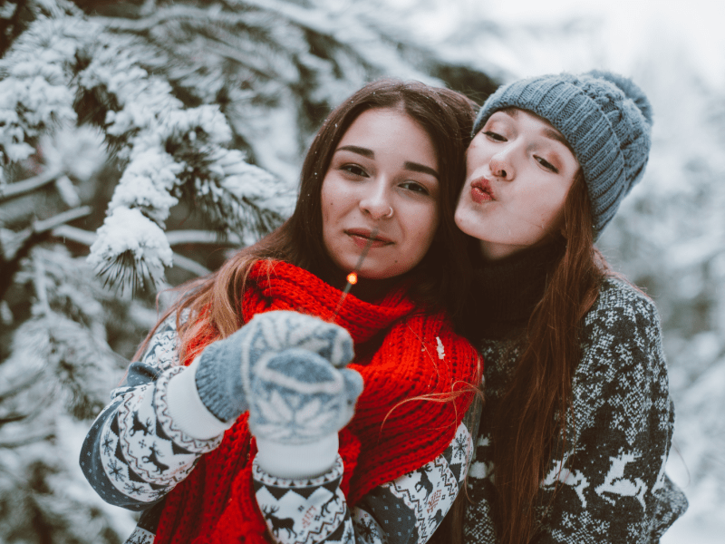 Zwei Frauen vor einer Winterlandschaft, mit einer Wunderkerze in der Hand, die sich umarmen und glücklich in die Kamera schauen.