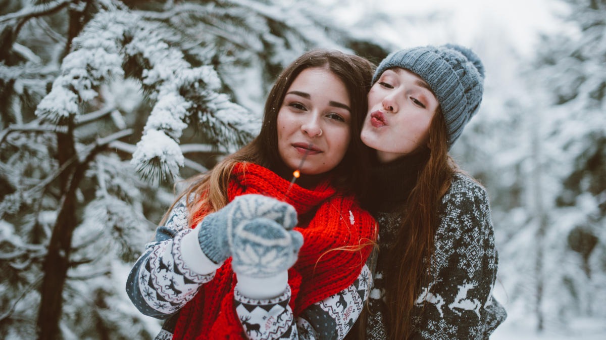 Zwei Frauen vor einer Winterlandschaft, mit einer Wunderkerze in der Hand, die sich umarmen und glücklich in die Kamera schauen.